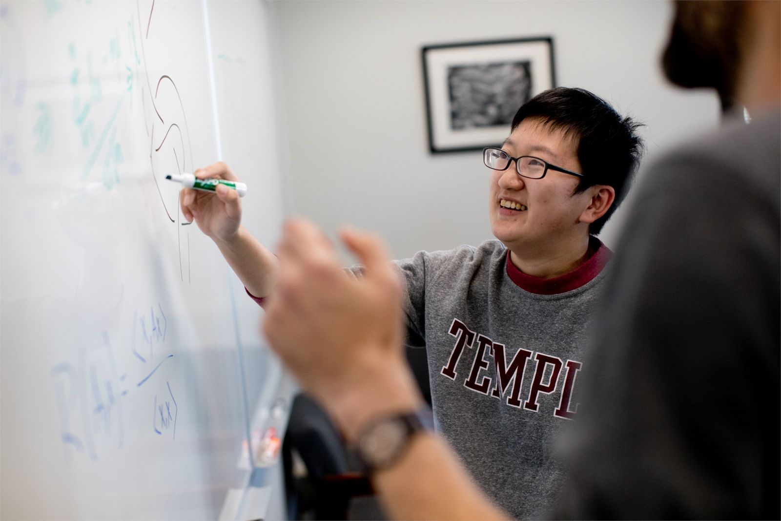 Student writing out math equation on white board with College of Science and Technology professor looking on in a Temple classroom. 