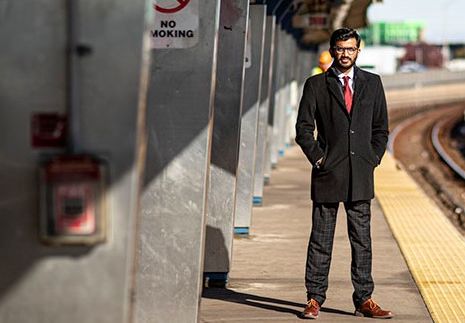 A man in a suit stands with his hands in his pockets on an elevated train platform.