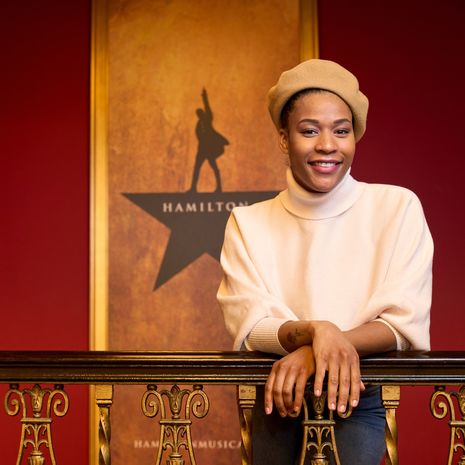 A woman wearing a tan beret and a beige sweater leans on a railing inside the Hamilton theater.