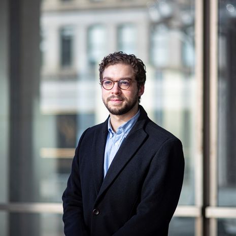 A man with a beard and wearing a suit poses in front of a window.