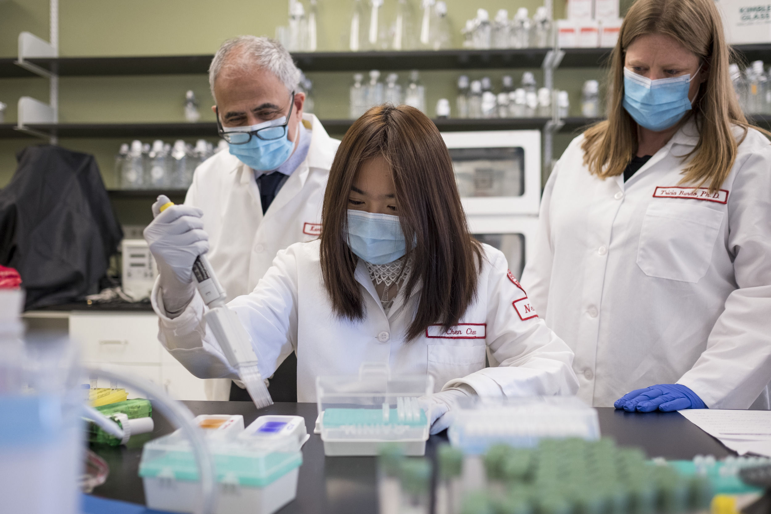 Three researchers wearing white lab coats and wearing masks work together in the lab