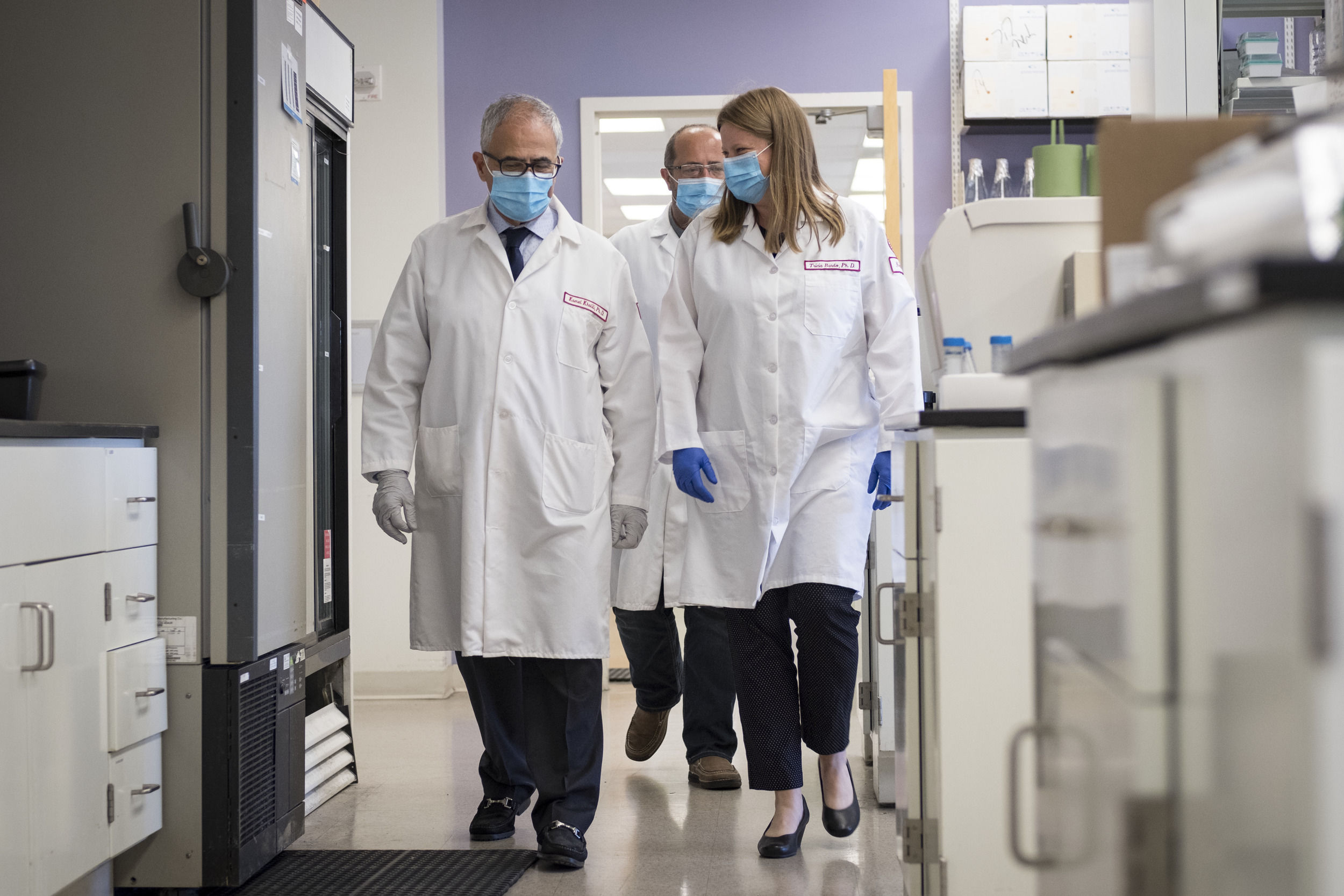 Three people wearing white lab coats and masks walk through the HIV research lab