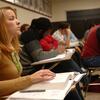 A classroom of students sitting at desks with a woman in the forefront looking towards the front.