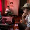 Two students sitting on set working on their laptops.