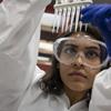 A student wearing a white lab coat and goggles inspects some test tubes.