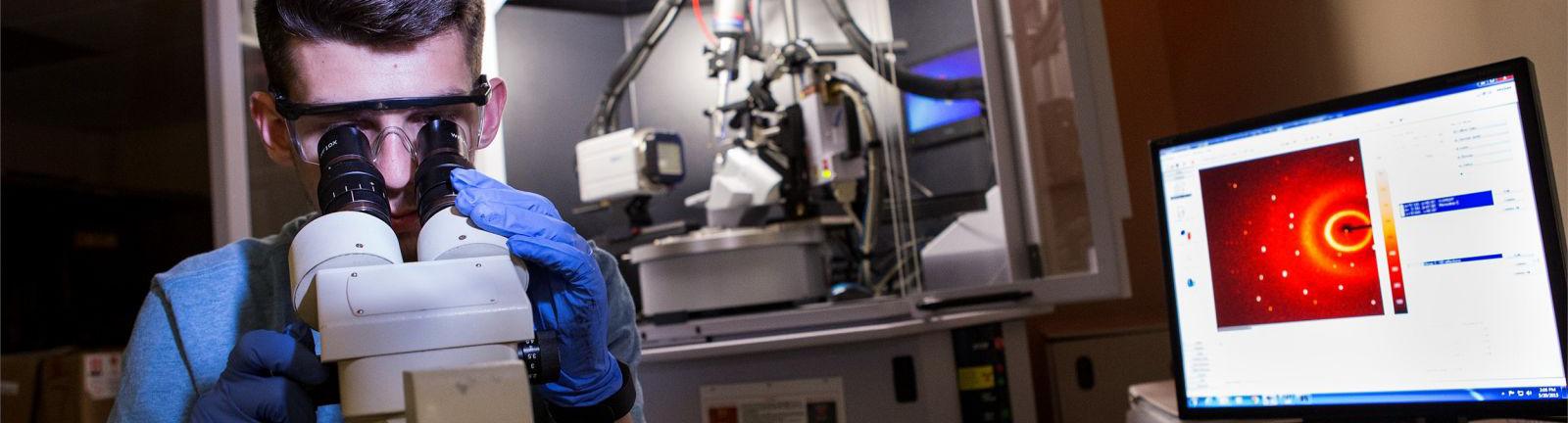 Chemistry student looking through a microscope in a lab surrounded by computer and equipment.