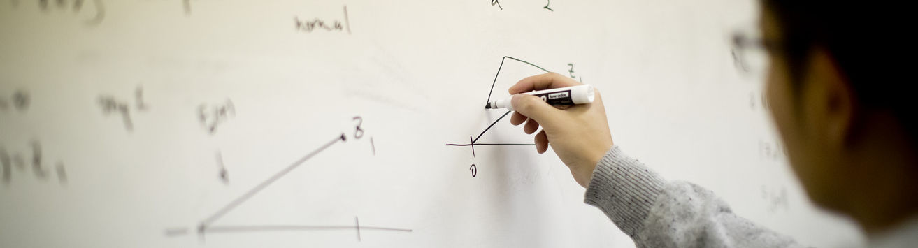 A student solves a math problem at a whiteboard in a classroom.