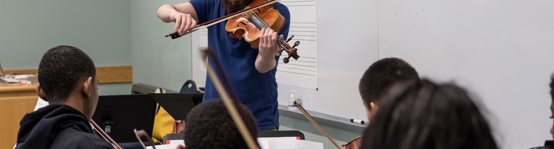 A violin teacher plays at the front of the class in front of young students.