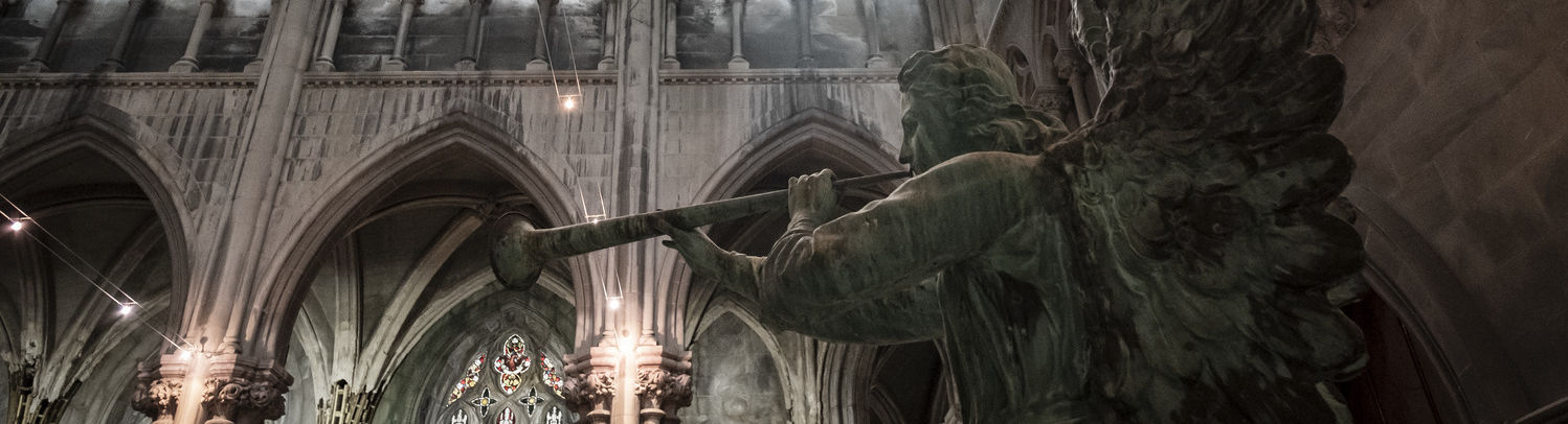 A sculpture of an angel with a trumpet is shown in front of a vaulted ceiling and light-filled windows of a church.