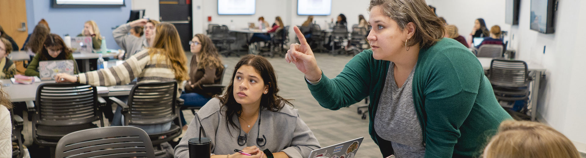 A professor points something out to a student in a classroom with students sitting at tables.