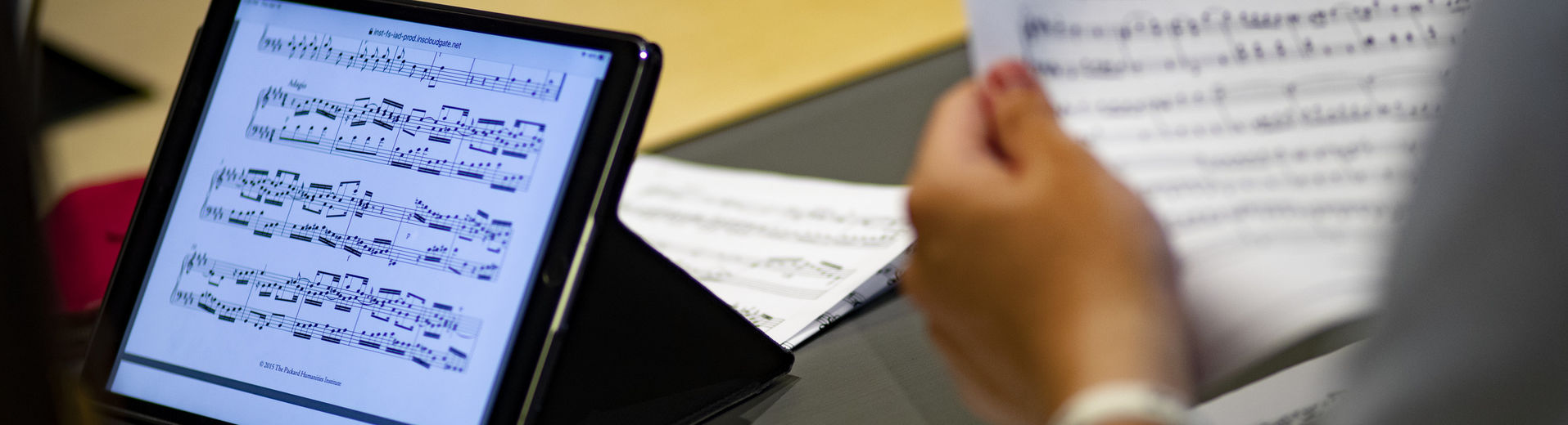 One student looks at a music score on a tablet while another student holds a paper score.