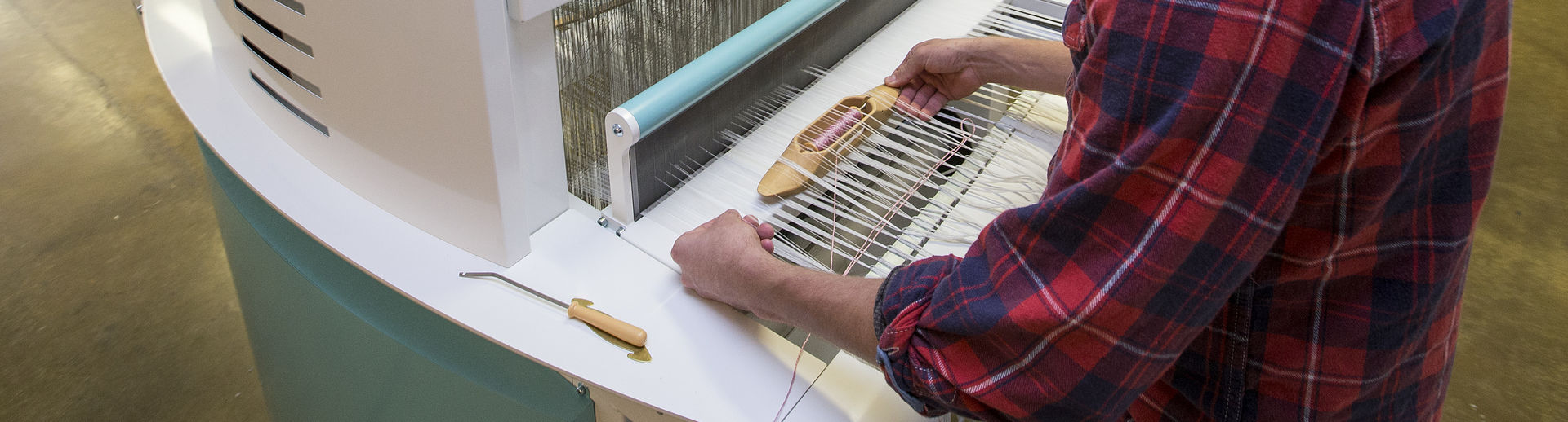 A student wearing a plaid shirt and yellow hat works on a loom.