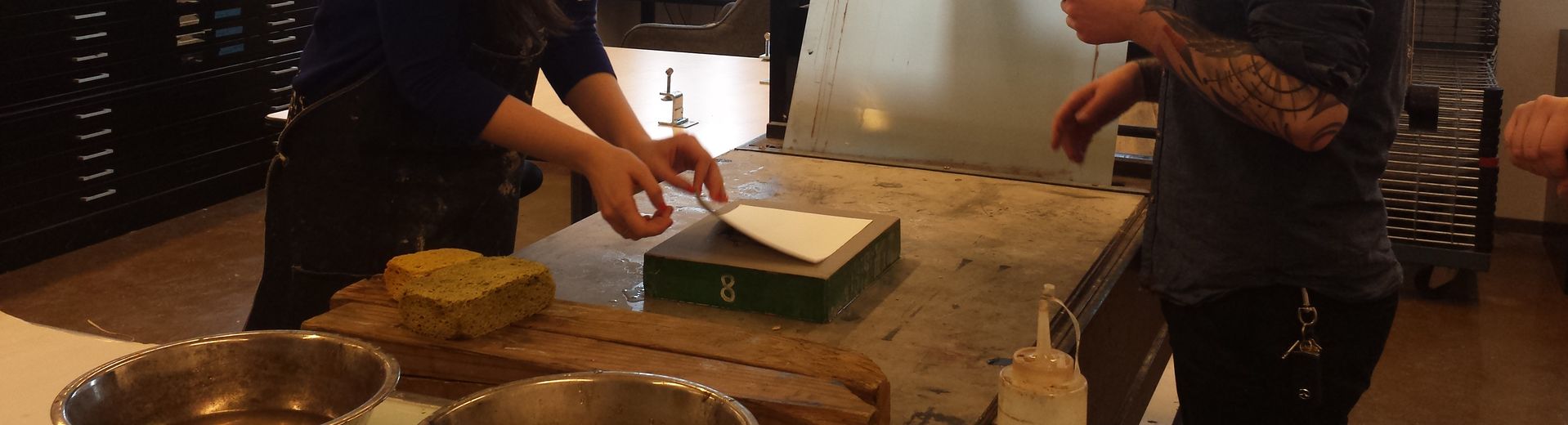 Two students look over a table where one student is peeling a sheet of paper from a block.