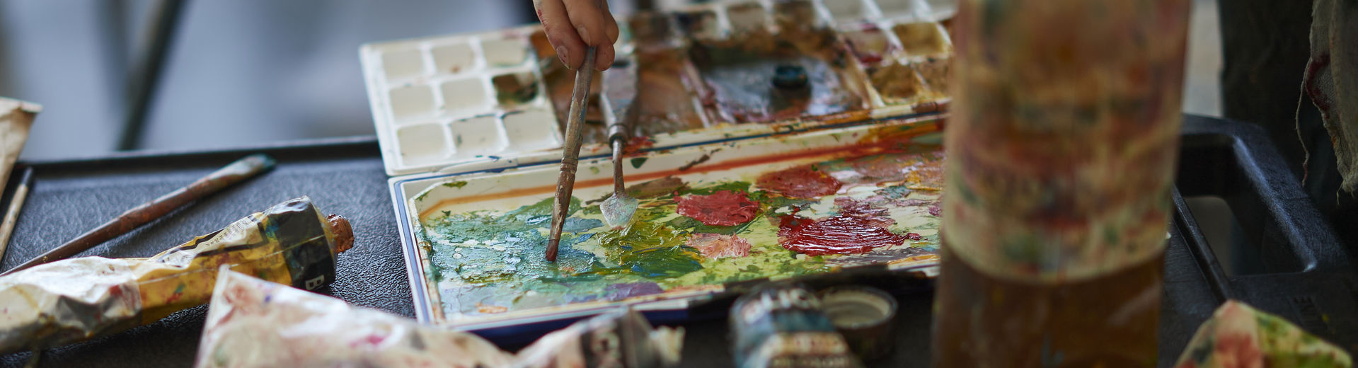 A hand holds a brush dipped into a palette of paint with tubes of paint all around the palette.