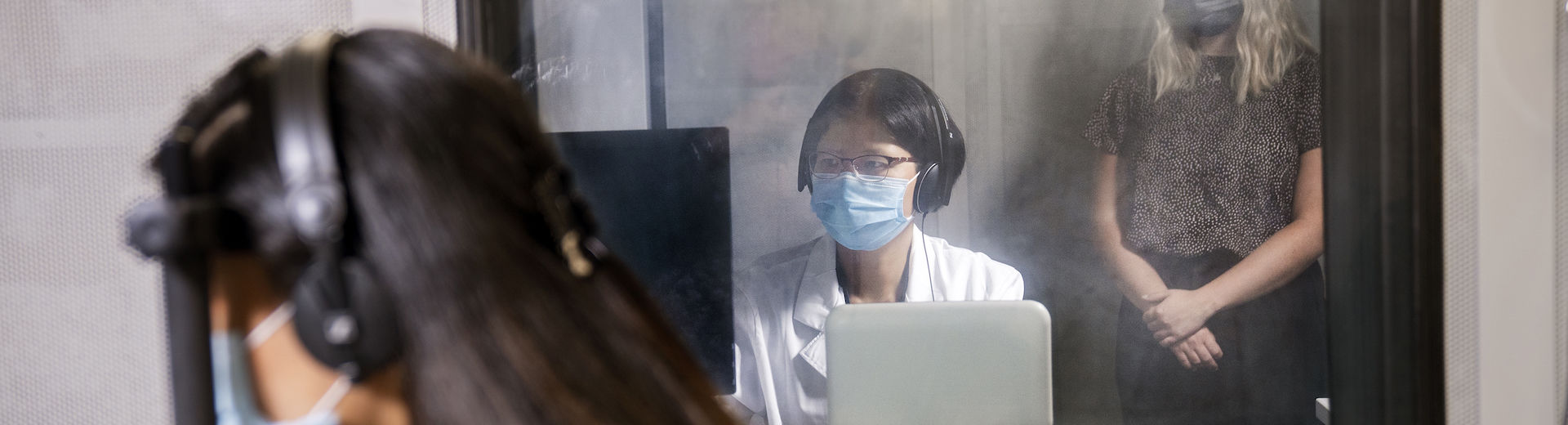 A masked student in a white shirt sits at a laptop with a woman standing behind here.