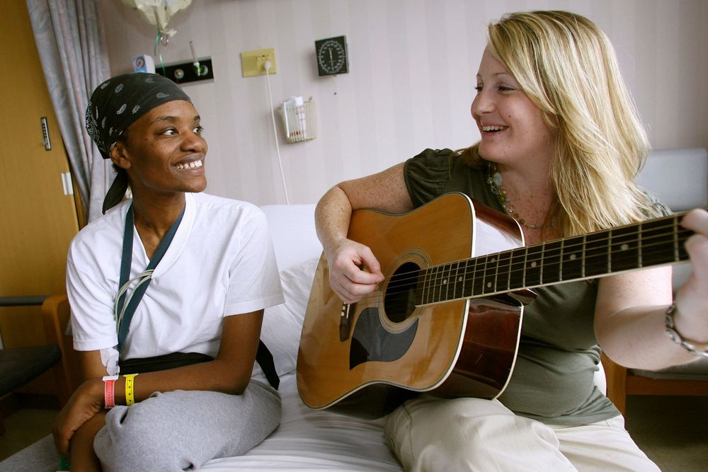 A Music Therapy student sits on a hospital bed next to a patient while playing the guitar.