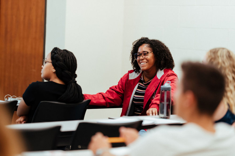 A female student in a red jacket smiles during a class discussion.