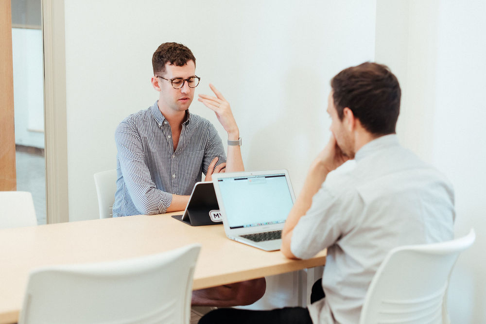 two students are engaged in a conversation in a study room with their laptops open in front of them.