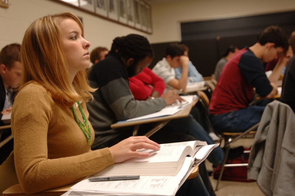 A classroom of students sitting at desks with a woman in the forefront looking towards the front.