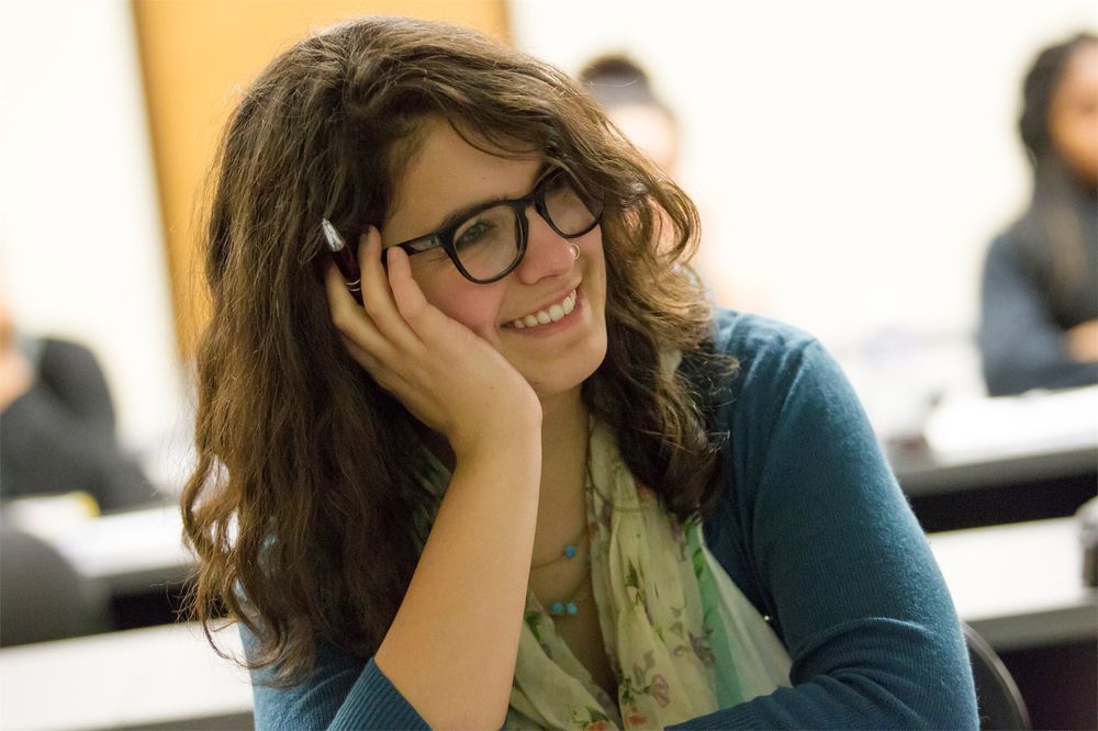 A bespectacled student, her face resting in her left hand, smiles during a class lecture.