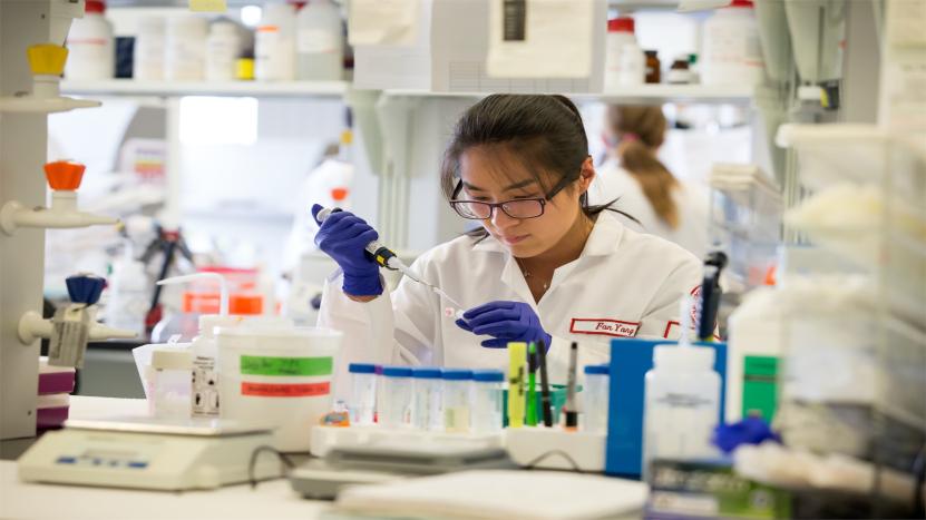 Female biology student wearing a lab coat and safety goggles performing an experiment in a research lab at Temple.