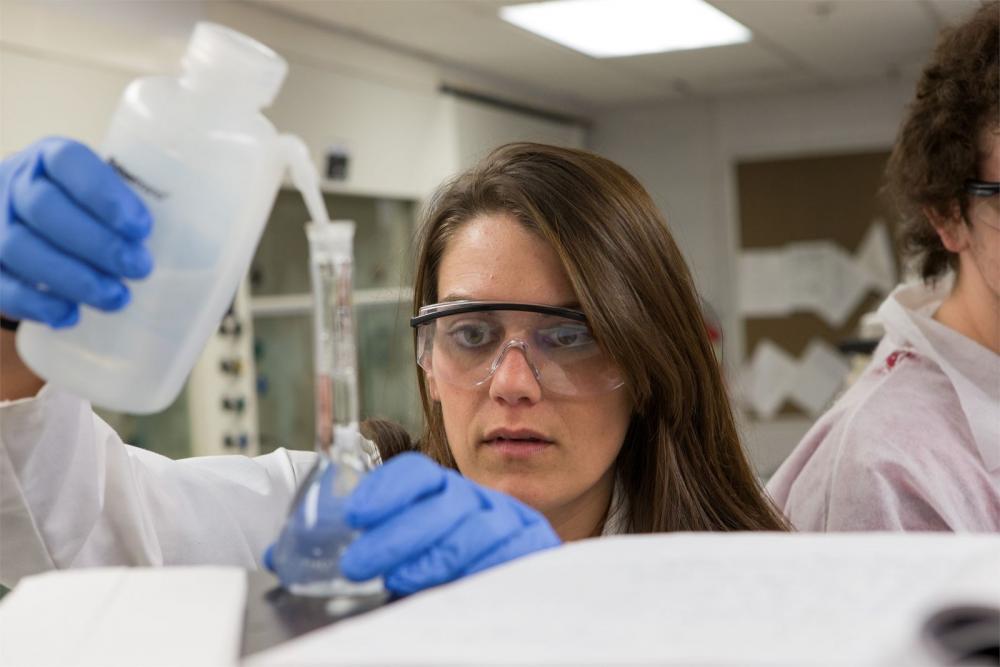 A Temple student wearing safety goggles and gloves pouring liquid into a beaker.
