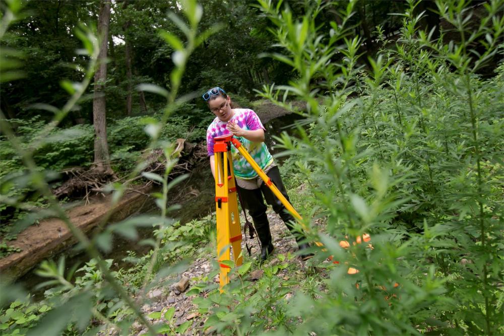 Temple student outside next to a riverbed using land surveying equipment.