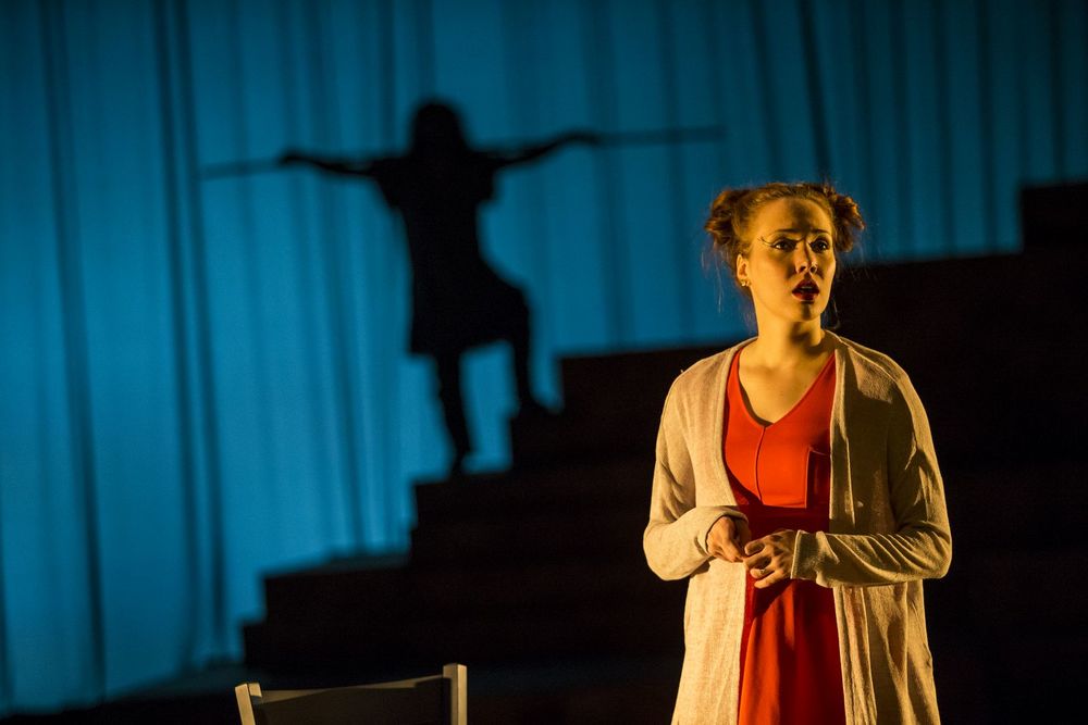 Women stands on stage with a shadow of a man behind her.