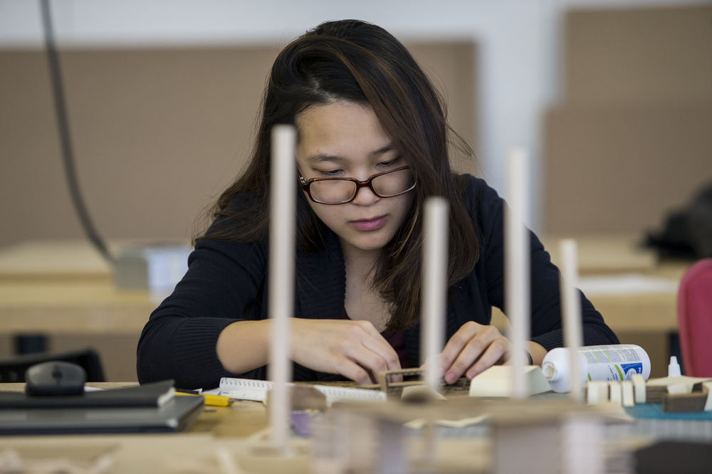 A woman with black hair and glasses works at a table with small pieces of cardboard.