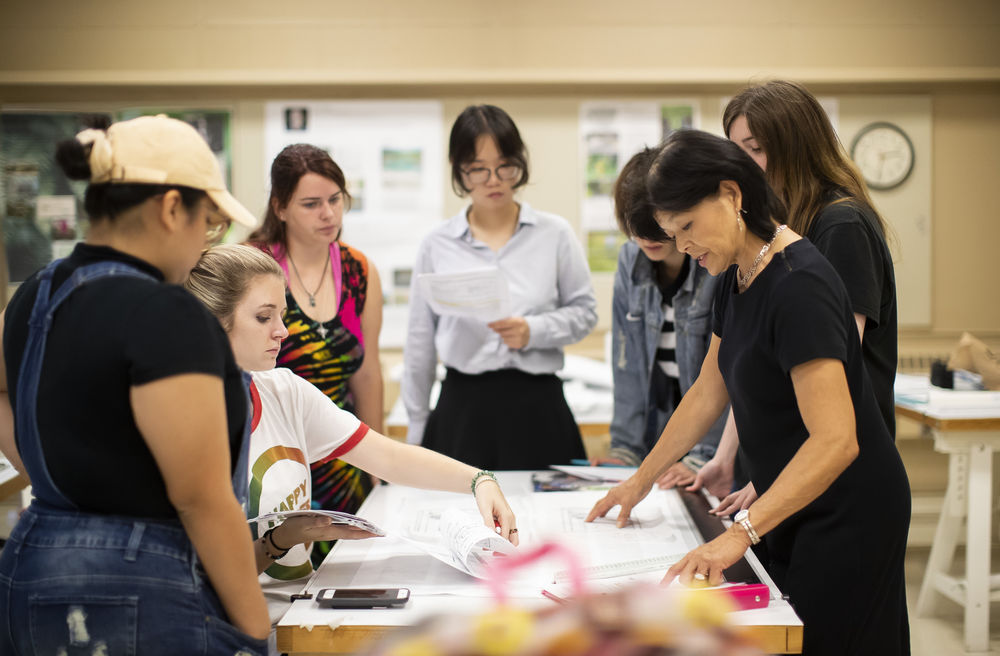 Students sit and stand around a table while a professor looks down at their work.