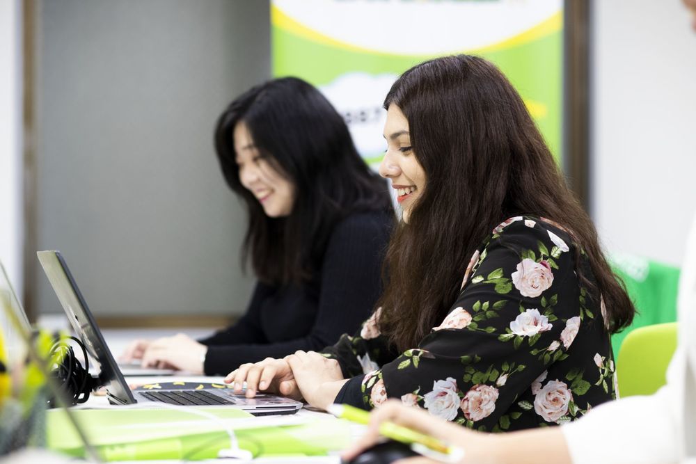 Two smiling women sit at a long desk looking at their laptops.