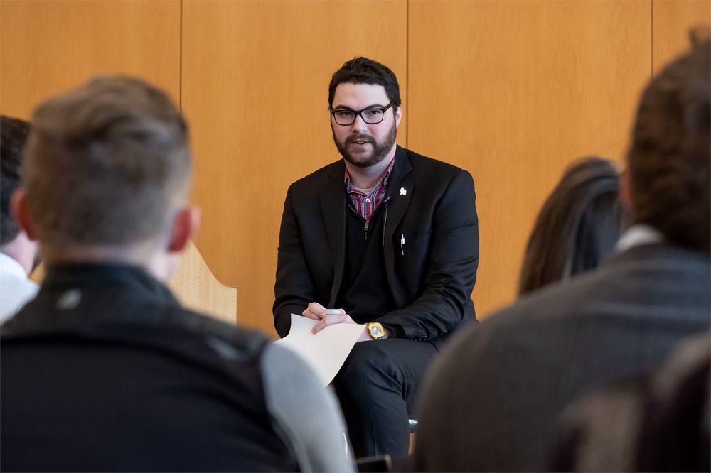 Bespectacled man sits on a stool and addresses a crowd.