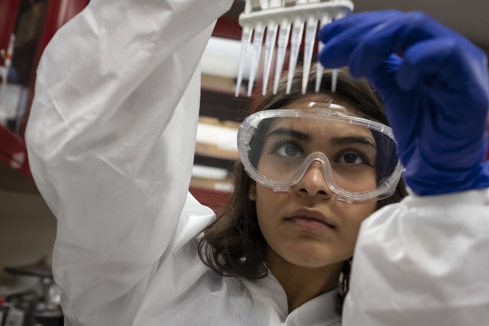 A student wearing a white lab coat and goggles inspects some test tubes.