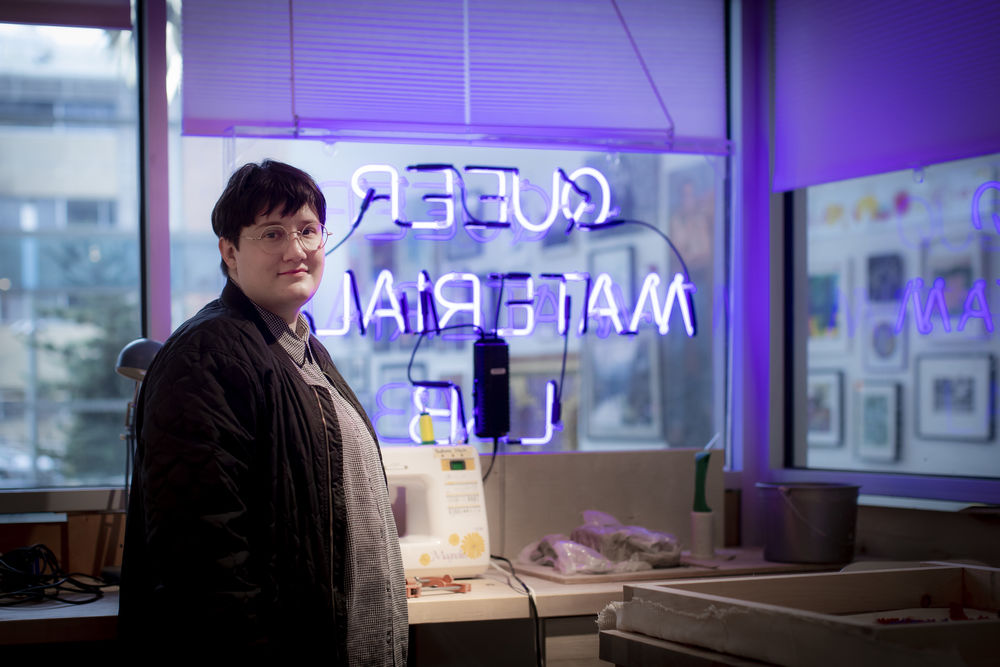 A person with glasses stands next to a sewing machine with a backwards neon sign behind them.