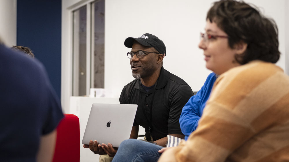 A man wearing a black baseball cap and holding his laptop sits with other students.