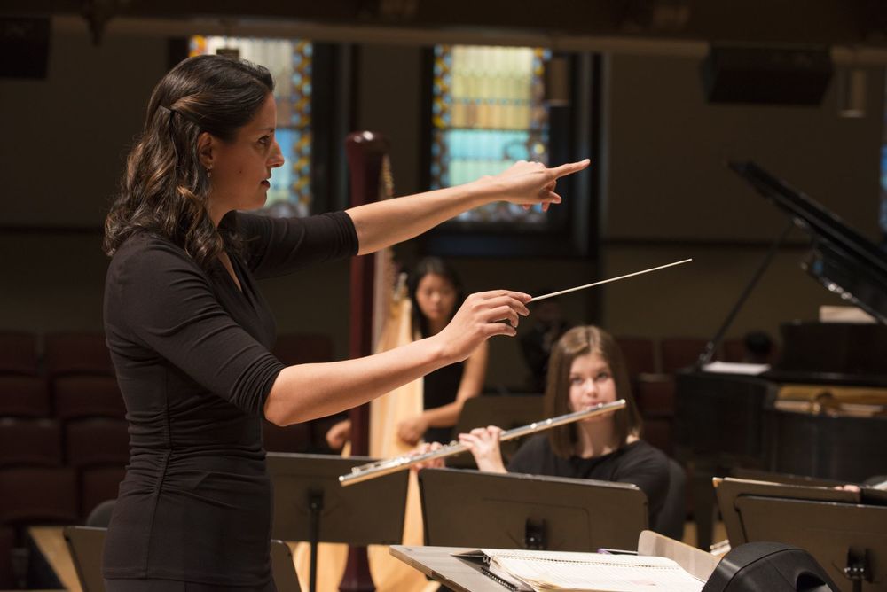 A woman conducts an orchestra with students playing flute and harp in the background.