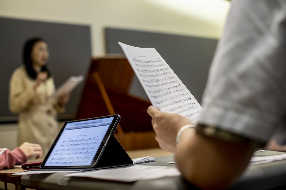 Music students looking at sheet music both on paper and on a computer while a professor lectures at the front of the classroom.