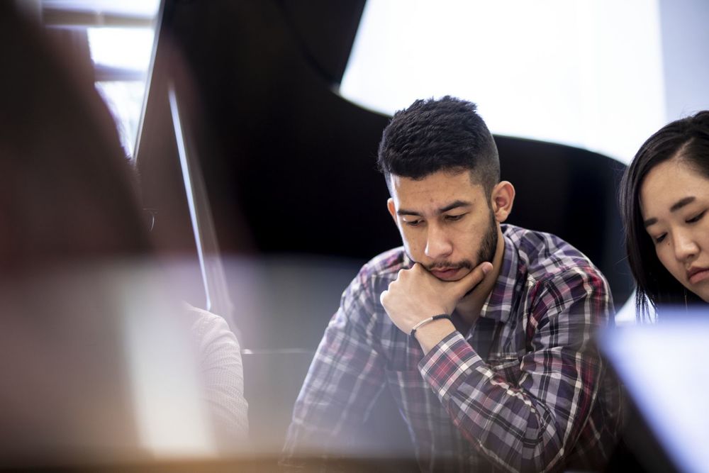 A Music Theory student, with hand on chin, looks down at his work while sitting among other students.