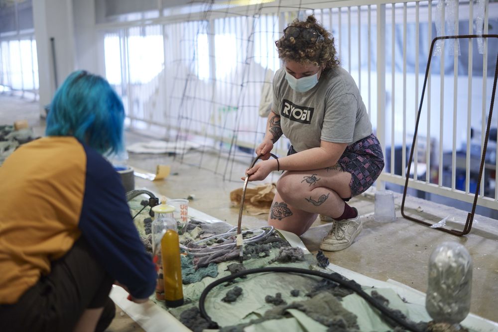 Two students bending on their knees work on a long textural sculpture on the ground.