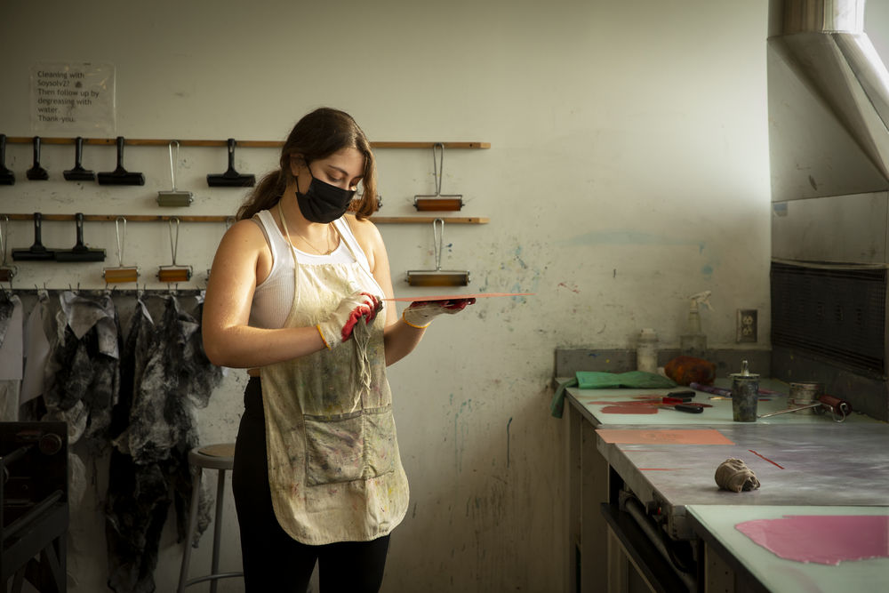 A student wearing a mask and gloves holds a square printing plate in an art studio.