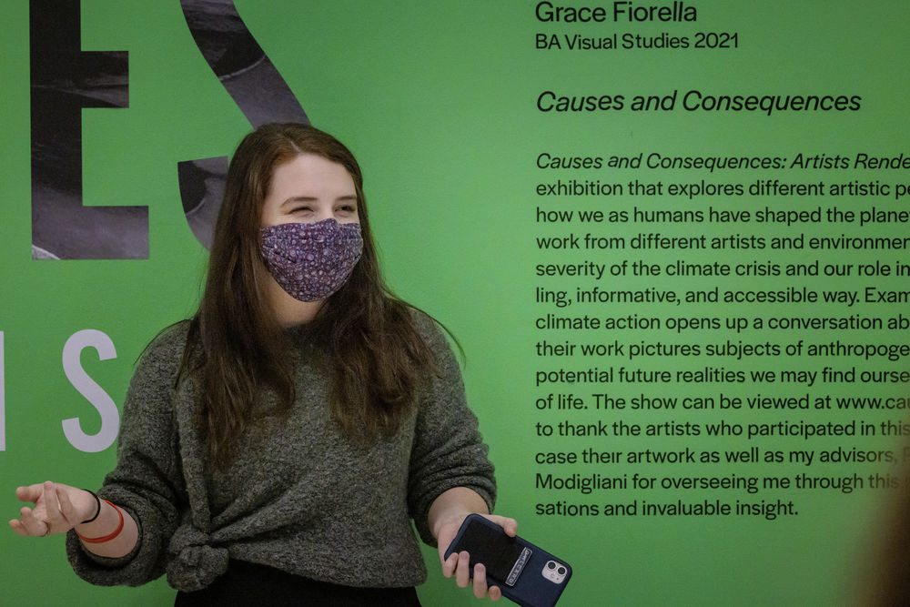 A masked Tyler student holds a phone as they stand in front of their work in a gallery.