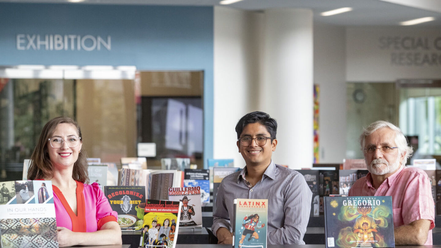 Professors Christina Baker, César Cabezas and Augusto Lorenzino standing in front of books on display in Charles Library for Hispanic Heritage Month